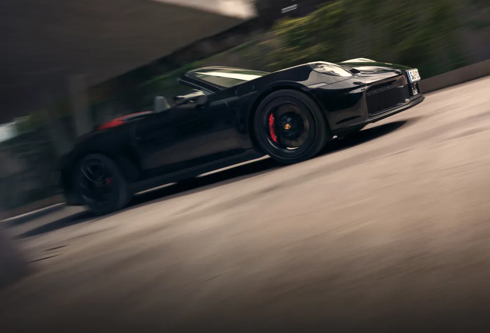 Porsche 911 Carrera GTS Cabriolet in Schwarz mit rotem Verdeck fährt durch Stadt. Blick auf die Seite.