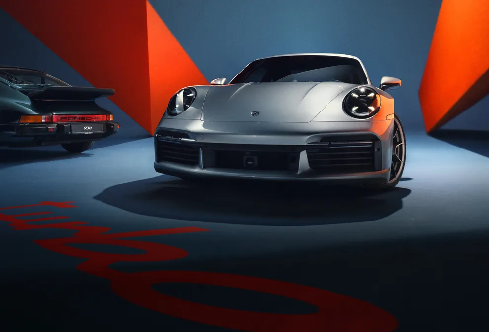 De Porsche 911 Turbo 50 Years Edition in GT-zilver metallic is van de voorkant te zien tegen een blauwe achtergrond. 