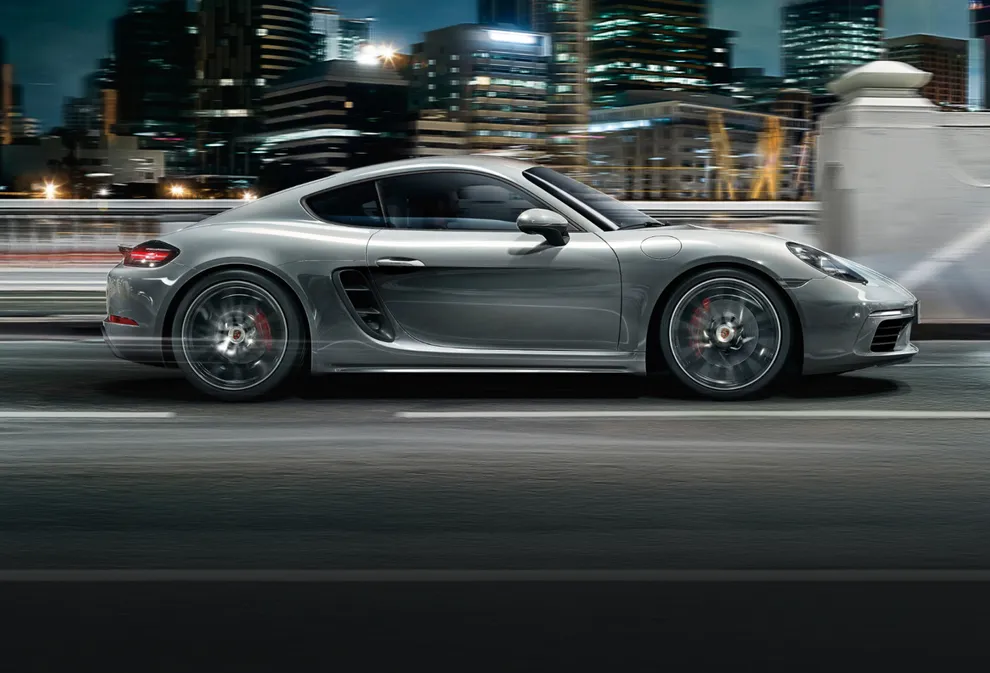 Porsche 718 Cayman S in GT-Silbermetallic fährt auf Straße vor Skyline einer Stadt. Blick auf Seite mit Flyline.