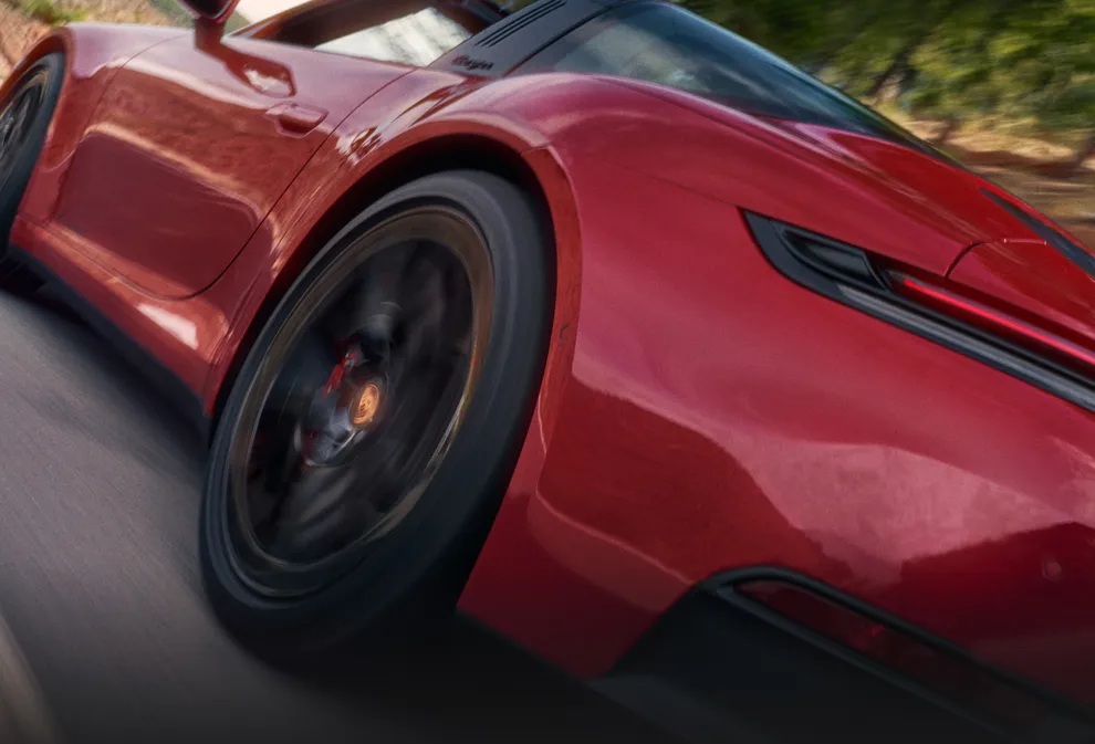 Porsche 911 Targa 4 GTS in Carmine Red driving on a road through a mountainous landscape. View of the side and rear end from behind.