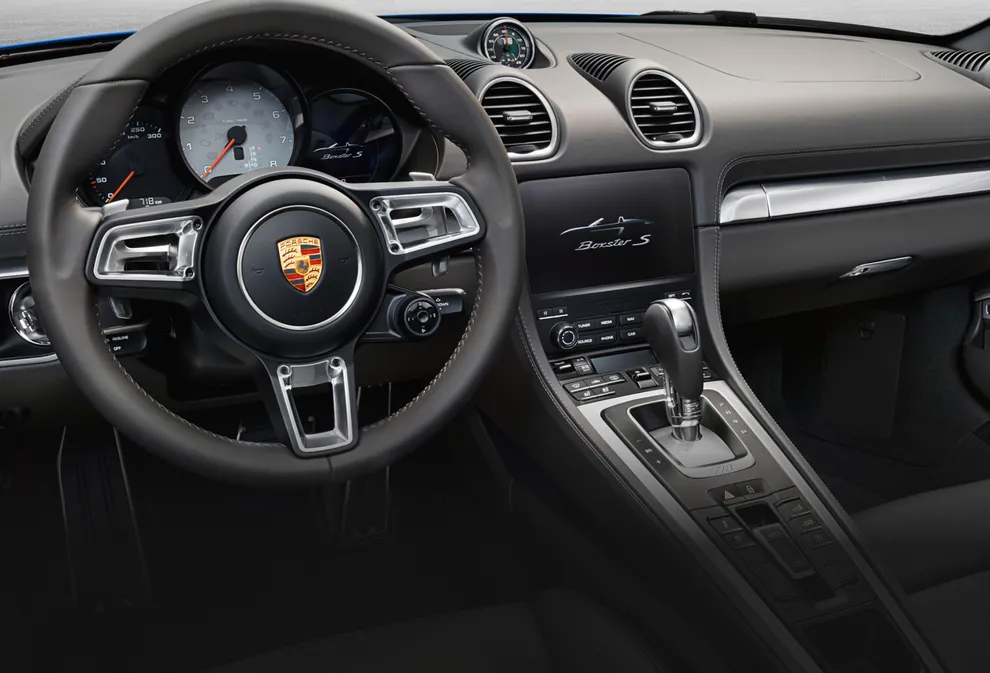 Interior del Porsche 718 Boxster S en color Gris Ágata. Vista del puesto de conducción centrada en la consola central y el volante.
