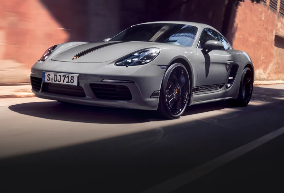 Porsche 718 Cayman Style Edition серого цвета (Arctic Grey) с пакетом контрастной отделки черного цвета едет по населенному пункту. Вид спереди на переднюю и боковую части.