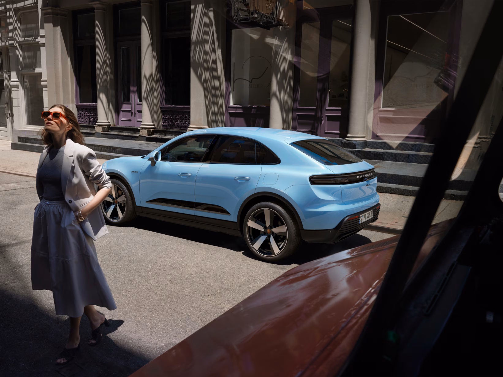 Porsche Macan in Frozenbluemetallicsteht in Stadt am Straßenrand. Blick auf die Seite. Im Vordergrund geht eine Frau vorbei.