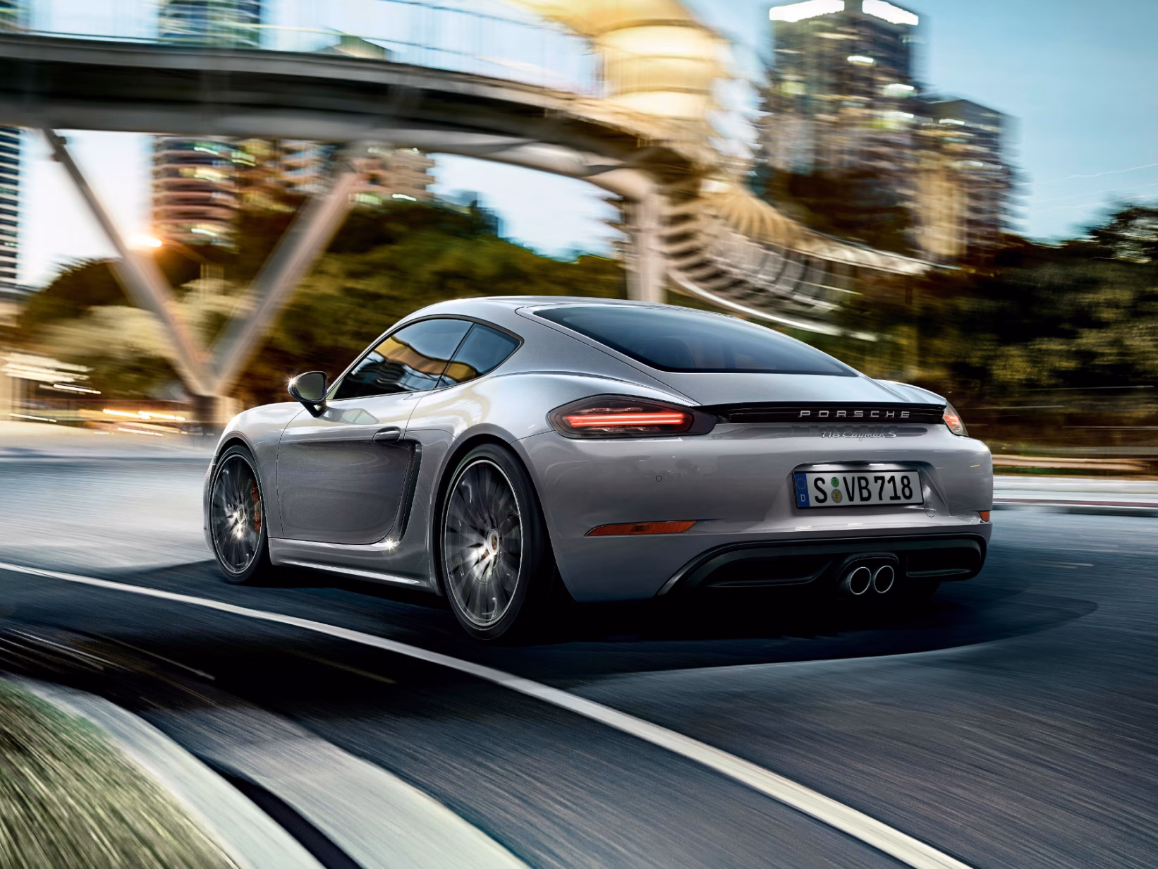 道路を走行するGT シルバー メタリックのPorsche 718 Cayman S、リアビューとサイドビュー