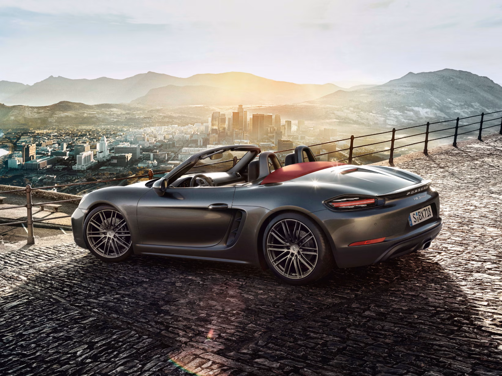 <p>Porsche 718 Boxster in Grigio Agata metallizzato parcheggiata in un punto panoramico di fronte a una catena montuosa. Vista laterale e posteriore.</p>