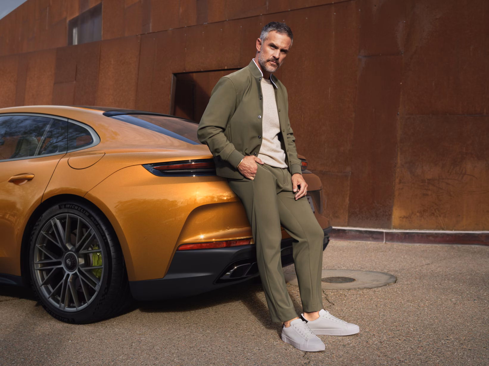 Porsche Panamera Turbo E-Hybrid in Madeira Gold Metallic in front of a rusty wall. View of the rear end. A man leaning on the rear end.