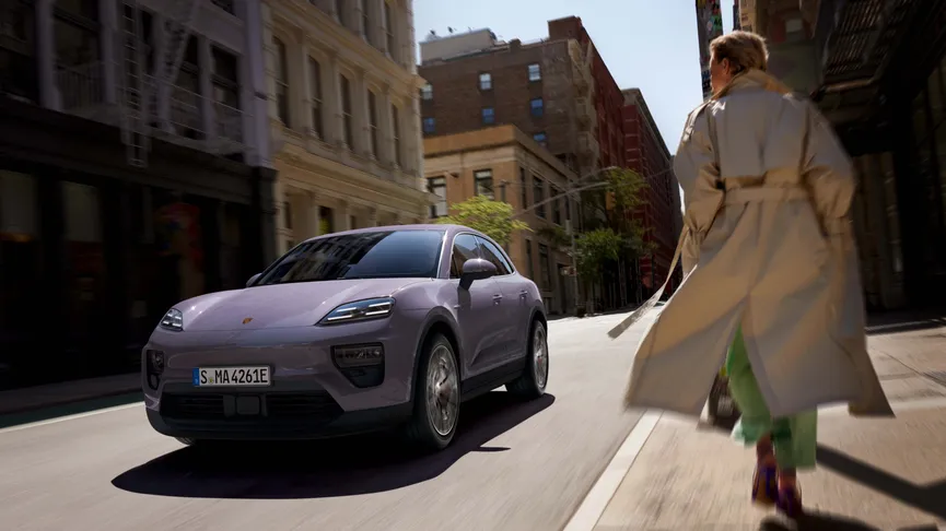 A purple SUV drives on a city street as a person in a trench coat walks on the sidewalk. Urban buildings line the background.