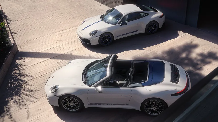 A high-angle shot shows two white Porsche 911 Carrera T, parked outside a modern house with the shade of a palm tree hanging over it. 