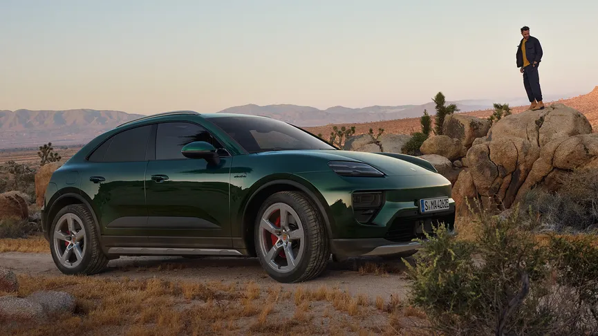 A dark green Porsche Macan 4S parked on a dirt road in a desert landscape. A man in a dark jacket is standing on a rocky outcrop in the background. A tall, thin palm tree is in the foreground.