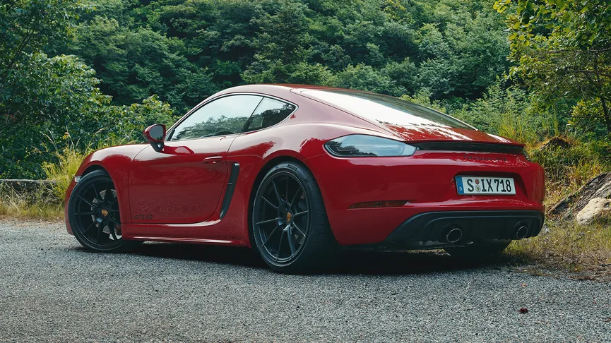 A low, rear 3/4 view of a red Porsche 718 Cayman GTS 4.0 parked on a gravel shoulder next to a stone wall. Lush green trees and hills rise in the background under a bright sky.