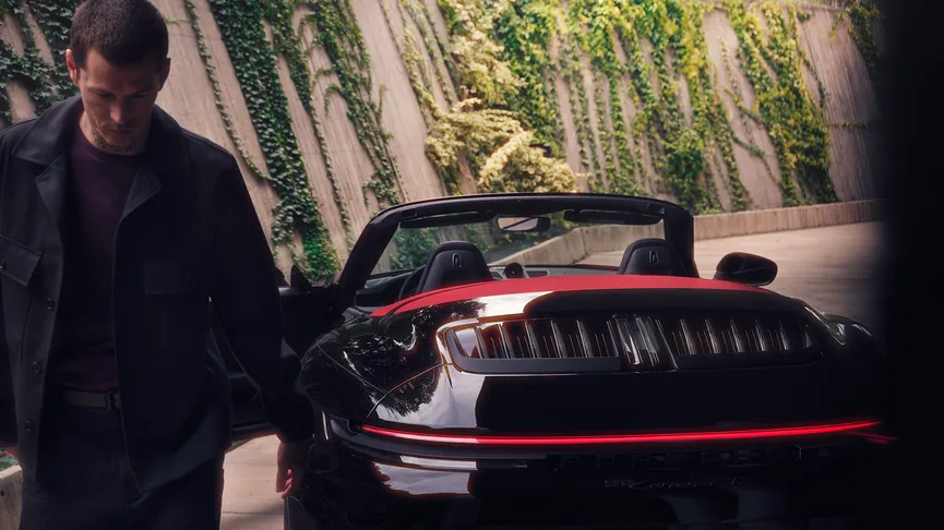 A dark, moody shot shows a man in a black jacket and jeans walking away from a black Porsche 911 cabriolet. The car's taillights are illuminated, and its red interior is visible