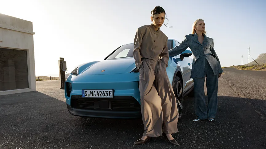 A light blue Porsche Macan Electric parked on a road next to a concrete building. Two people are standing near the car. The background includes a road and a coastal landscape.