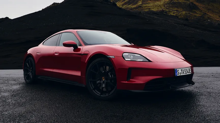 A red Porsche Taycan GTS is parked on a wet asphalt surface, with a dark, mountainous landscape in the background.