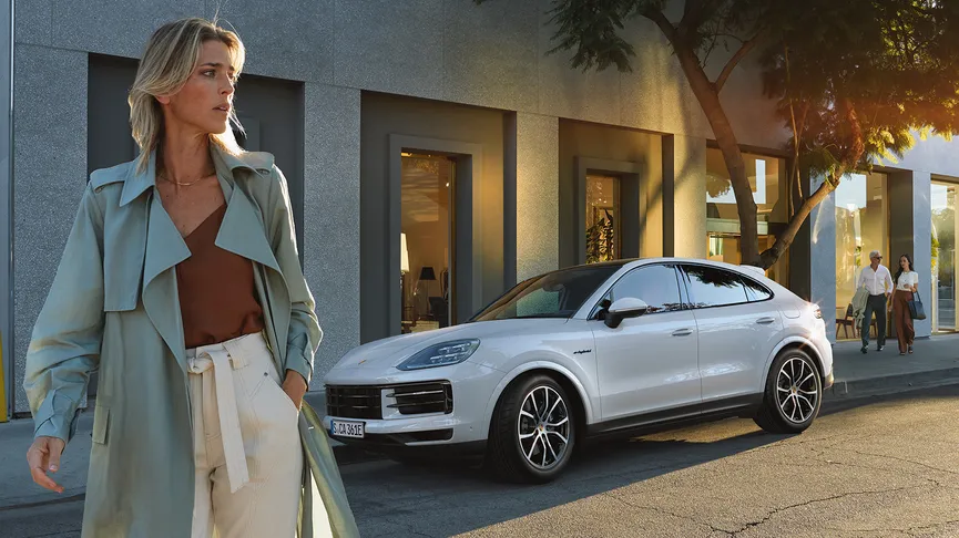 A white Porsche Cayenne Coupe parked on a city street next to a modern building with large windows. A person with blonde hair, wearing a light green trench coat, a brown top, and white pants, is walking towards the left in the foreground.