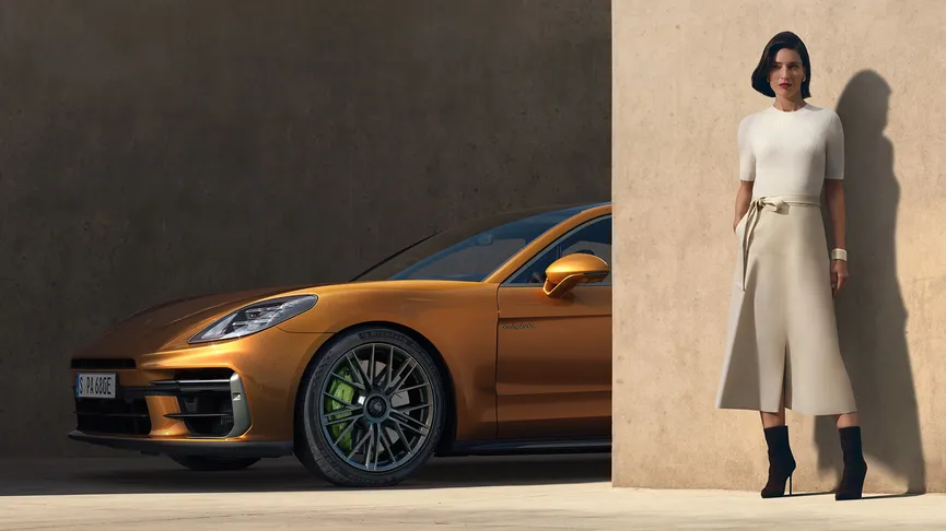 A woman in a cream-coloured top and skirt stands next to a golden-orange Porsche Panamera in a garage-like setting. The woman stands to the right, leaning against a tan-coloured wall.