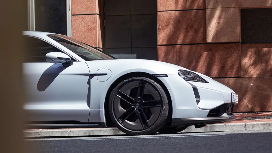 White Porsche Taycan, angled right with black wheels, parked on asphalt. A shaded reddish-brown building with a dark door and window is behind it. Bright sunlight. Blurred foreground.