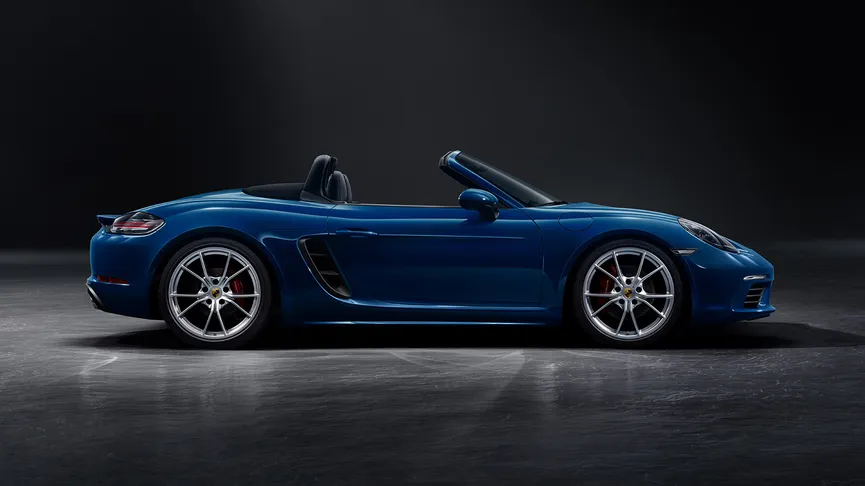 Side view of a blue Porsche 718 Boxster convertible with its black soft top down, on a dark background.