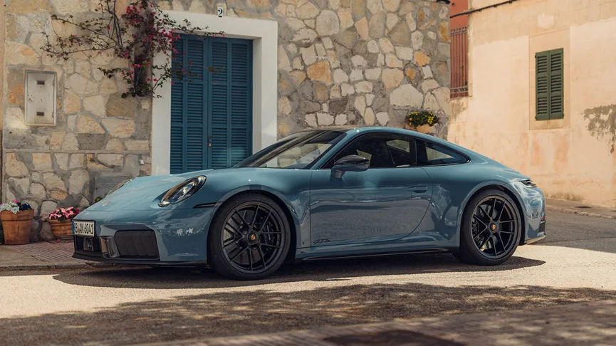 A Porsche 911 parked on a street in Mallorca. The car is parked on a paved street. The background includes buildings with stone and stucco walls, with a tree partially obscuring the view.