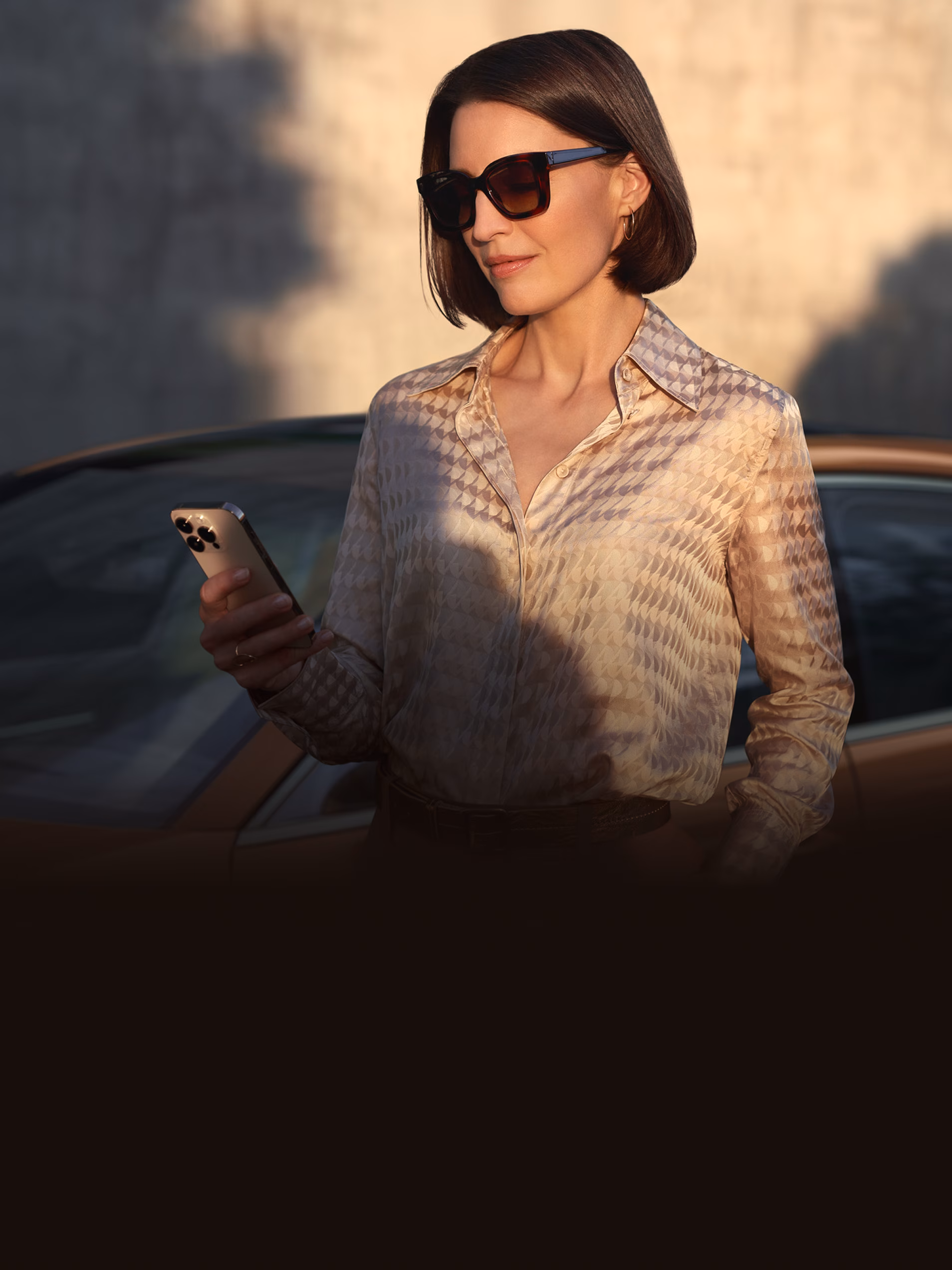 Una persona mira su smartphone con la app Porsche Connect. Al fondo, un Porsche Turbo E-Hybrid en color Madeiragoldmetallic.