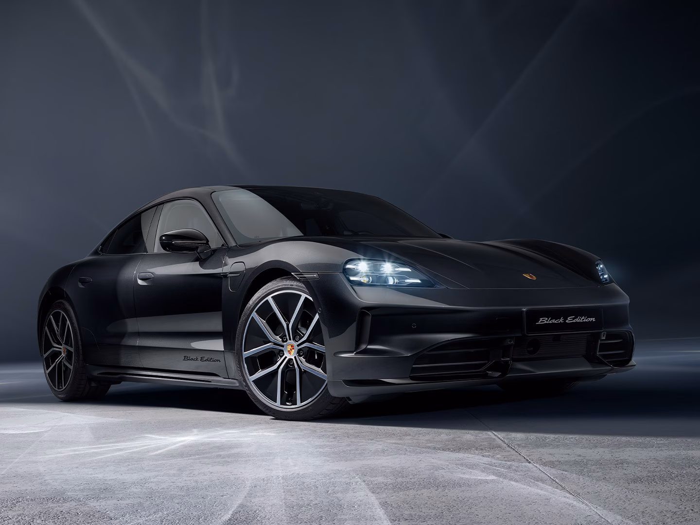 A sleek, black Porsche Taycan Black Edition electric sports car is parked in a dark, atmospheric studio. The car features "Black Edition" badging on the side skirt and front license plate area, complemented by large, multi-spoke alloy wheels with a silver and black finish. Dramatic, smoky light patterns swirl in the background and across the concrete floor, illuminated by the car's glowing LED headlights.