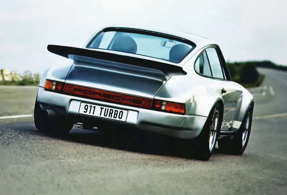 Er wordt een Porsche 911 RSR Turbo zijwaarts van achteren getoond die op een weg rijdt.