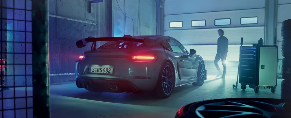 Porsche 718 Cayman GT4 RS in Arktikgrau mit Weissach-Paket steht in Garage. Blick auf Heck mit eingeschalteten Heckleuchten.