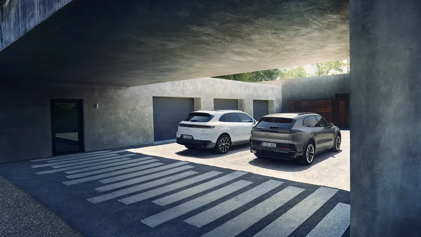 Two modern SUVs parked side-by-side in a minimalist concrete courtyard under a broad overhang, sunlight casting striped shadows on the ground.