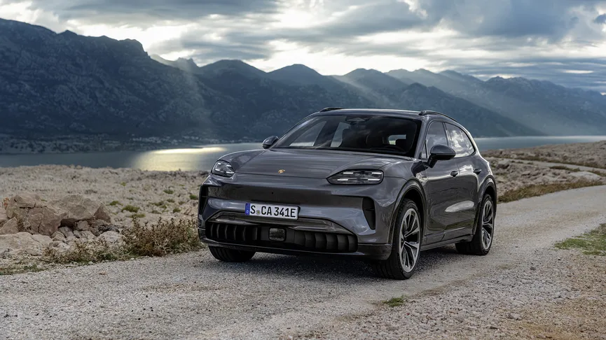 A sleek gray car parked on a gravel road with a mountainous landscape and sun rays breaking through clouds in the background.