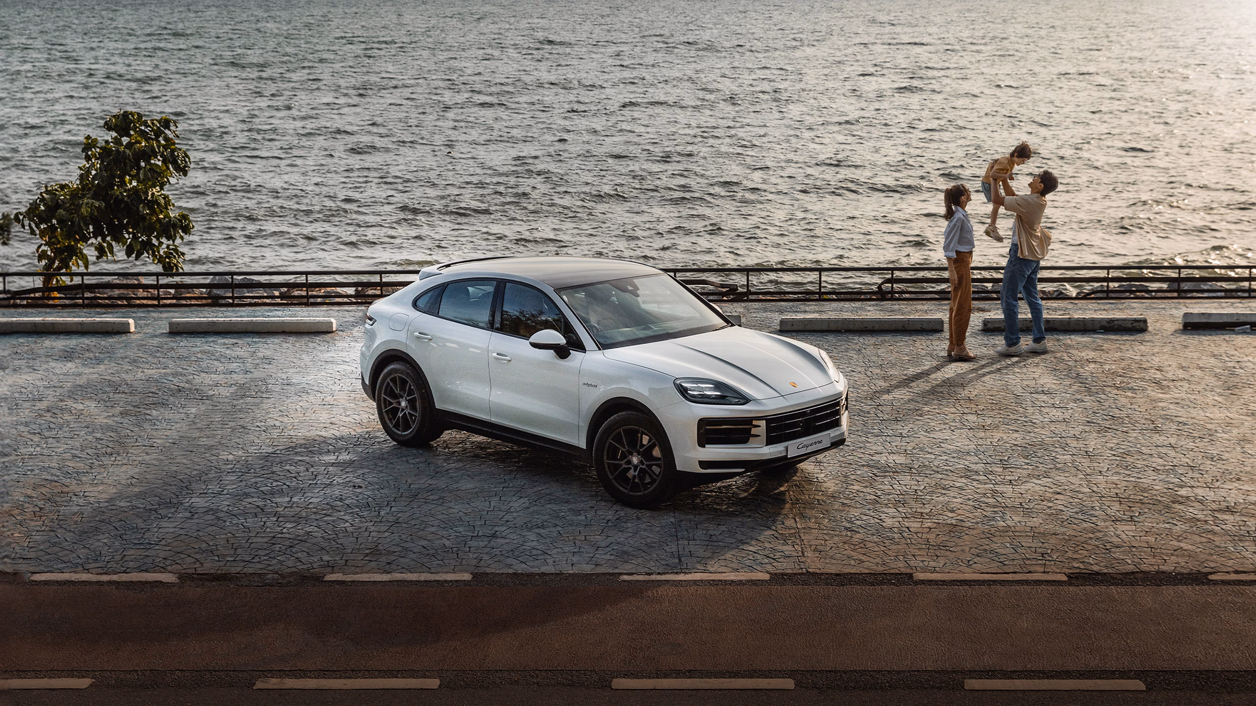 White Porsche Cayenne parked by a waterfront promenade, with a family playing in the background.