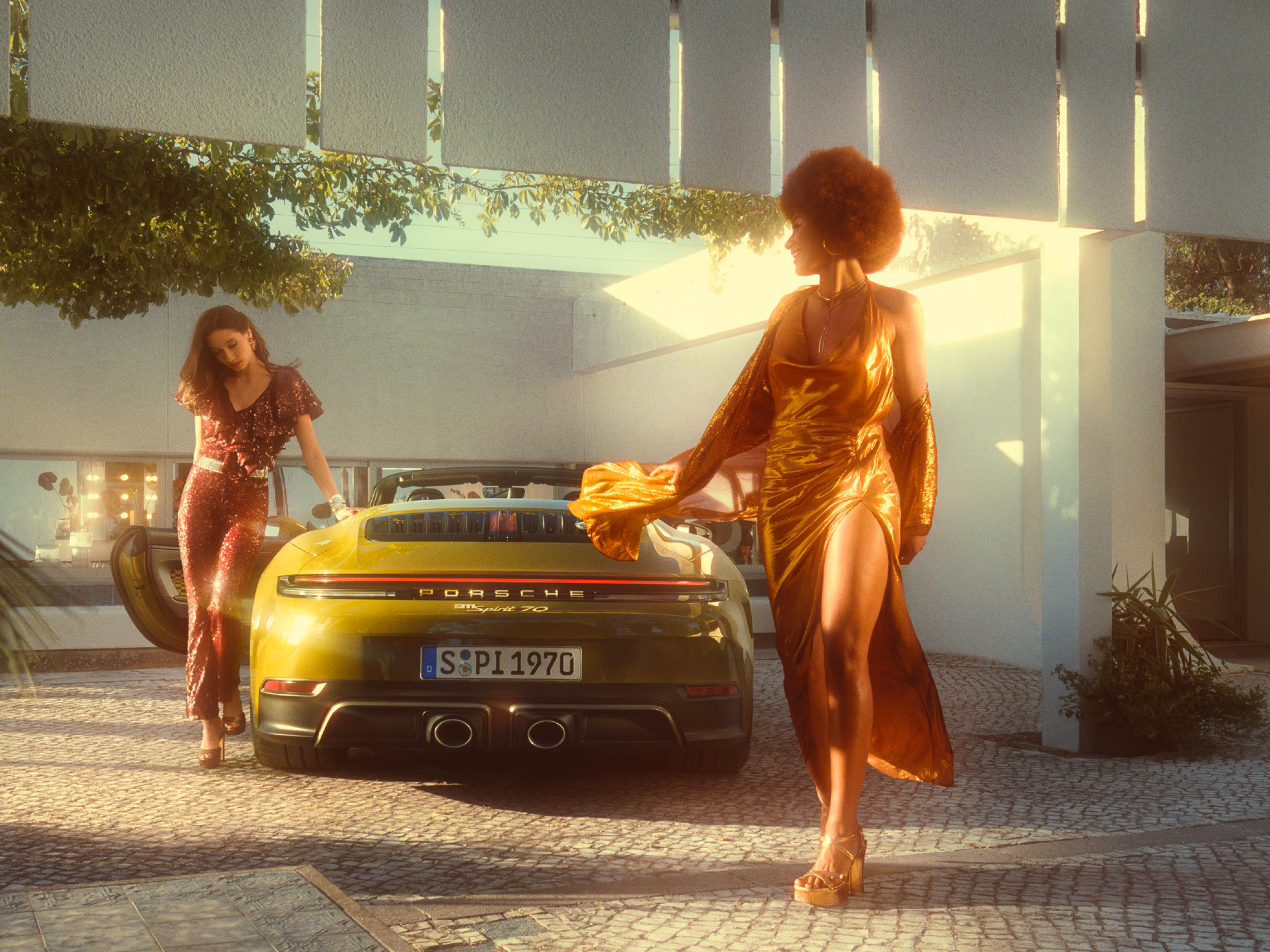 Porsche 911 Spirit 70 in an Olive Neo paint finish parked in a paved square. View of the rear end from behind. Two women wearing 70s clothing are getting out of the vehicle.