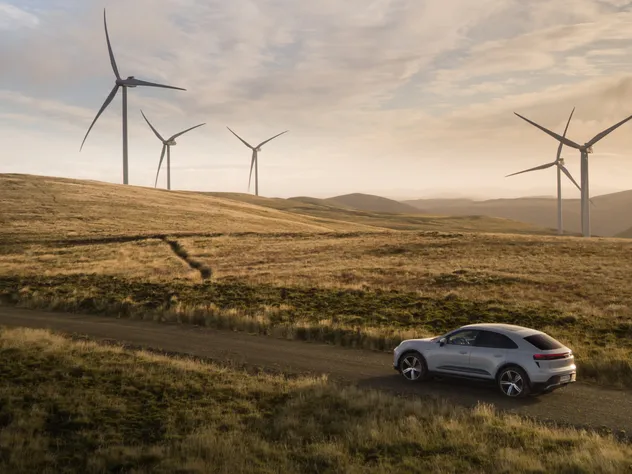 Un Porsche Macan Turbo Gris Glace Métallisé roule sur une route de campagne, traversant un paysage de prairies vallonnées. Des éoliennes se dressent en arrière-plan.