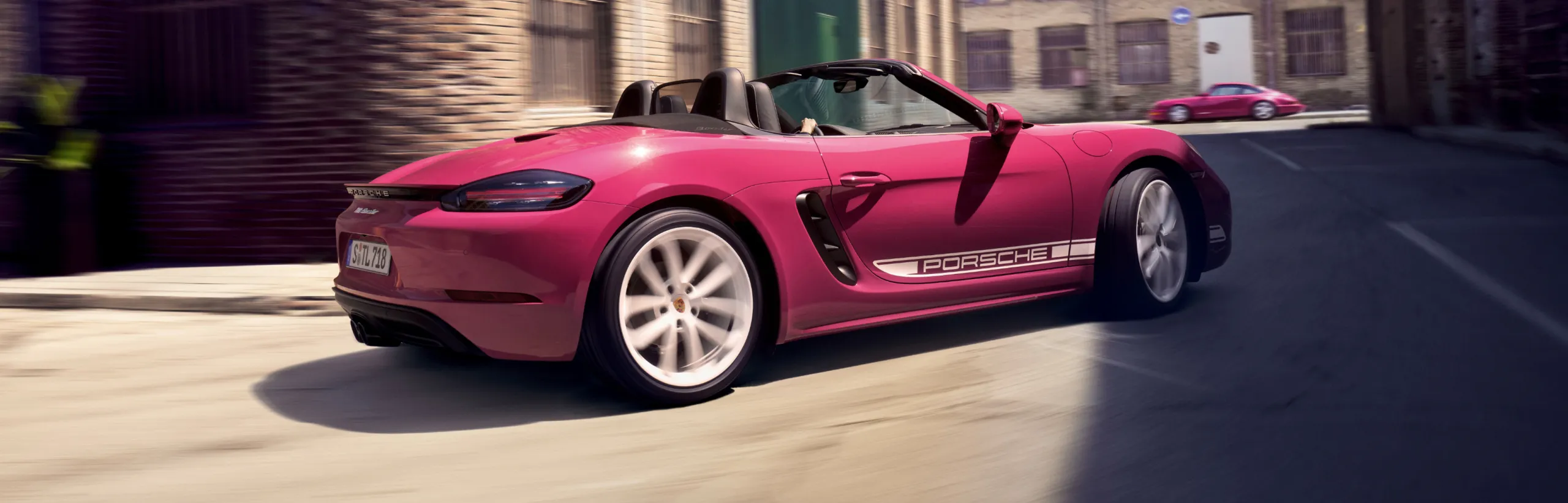Porsche 718 Boxster in Ruby Star Neo con pacchetto a contrasto bianco che affronta una curva. Vista della parte laterale e posteriore.