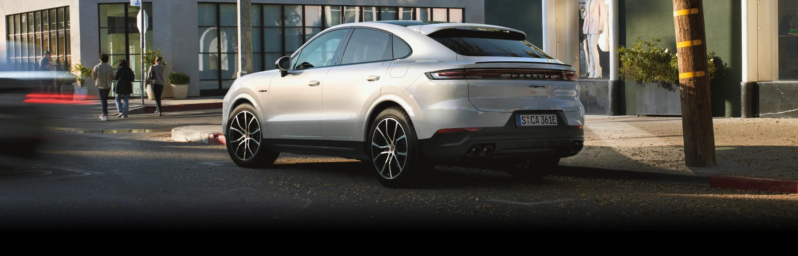 Porsche Cayenne E-Hybrid цвета белый металлик (Carrara White Metallic) едет по проселочной дороге, пролегающей на рельефной территории. Вид сзади и сбоку.