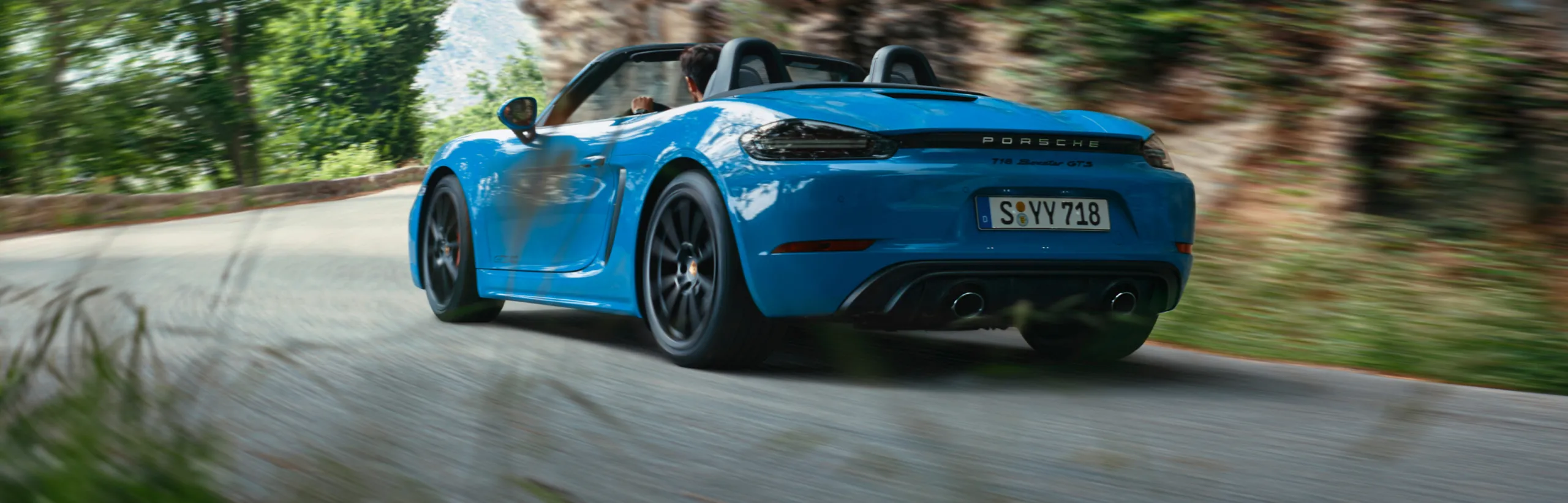 Porsche 718 Boxster GTS 4.0 en color Azul Shark circulando por una carretera de montaña. Vista de la parte trasera y el lateral.