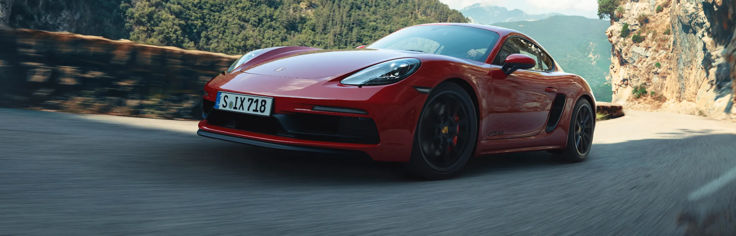 Porsche 718 Cayman GTS 4.0 en color Rojo Carmín circulando por una carretera de montaña. Vista de la parte delantera y el lateral.