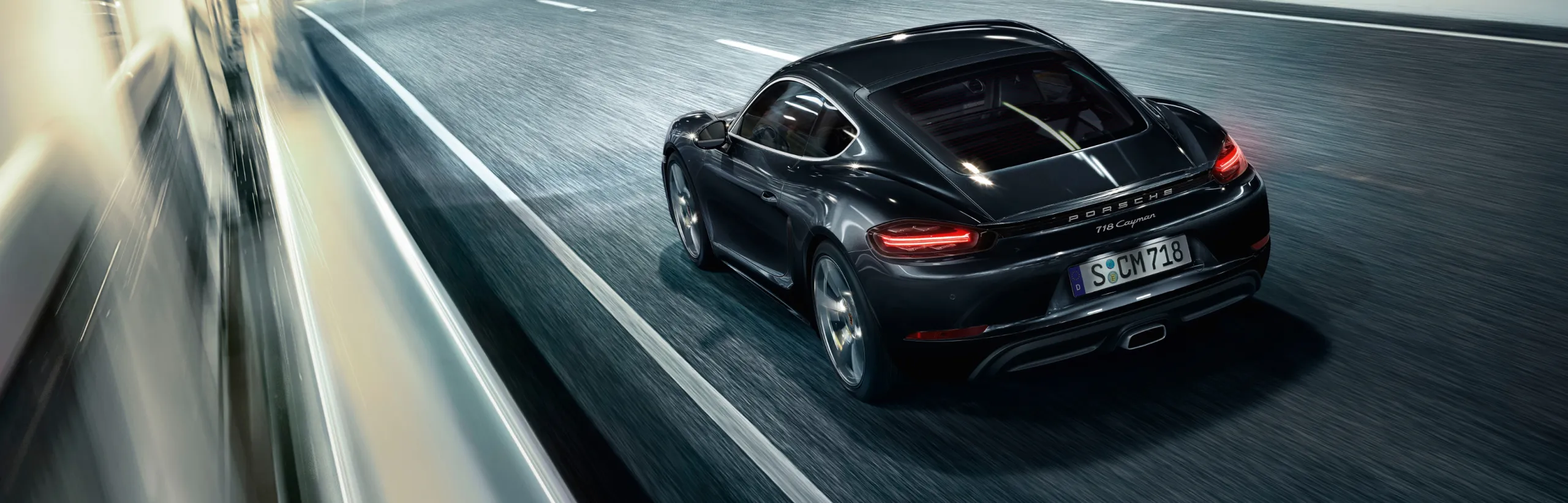 Porsche 718 Cayman цвета черный металлик (Jet Black Metallic) едет по скоростному шоссе в сумерках. Вид на заднюю и боковую части.