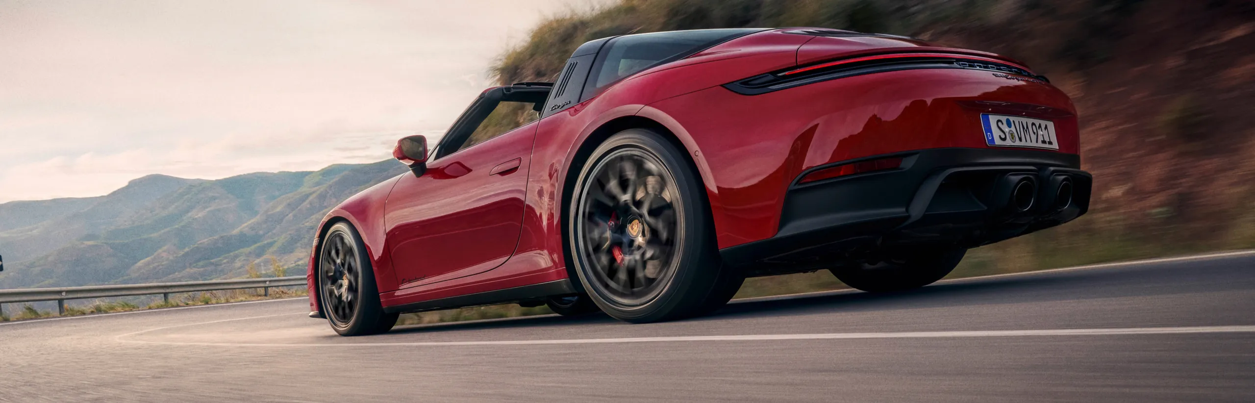 Porsche 911 Targa 4 GTS in Carmine Red driving on a road through a mountainous landscape. Rear and side view.