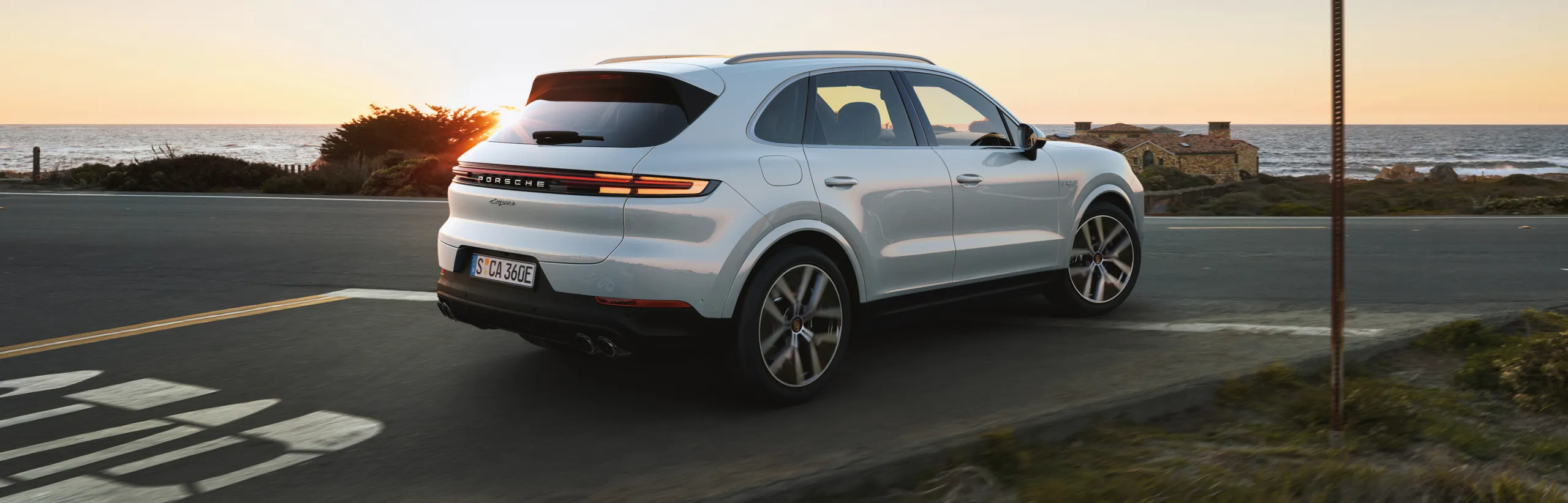 Porsche Cayenne E-Hybrid em Branco Carrara metálico passando por estradas rurais no interior do país. Vista da traseira e lateral.