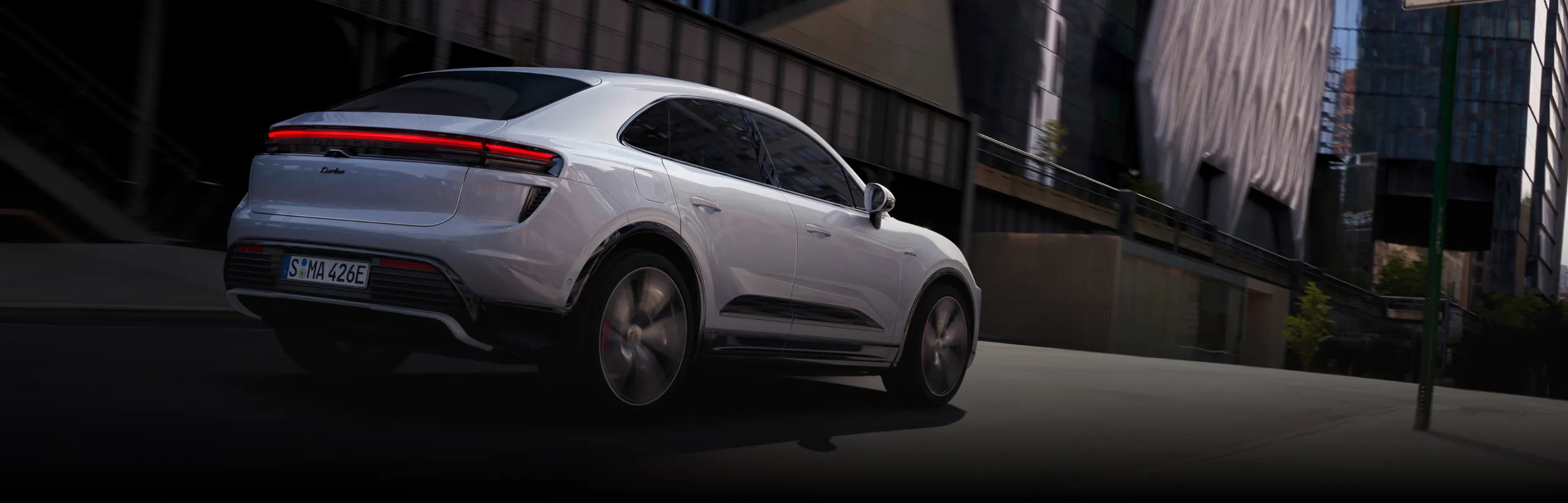 Porsche Macan Turbo in Ice Grey Metallic driving around a corner in a city. Rear and side visible.
