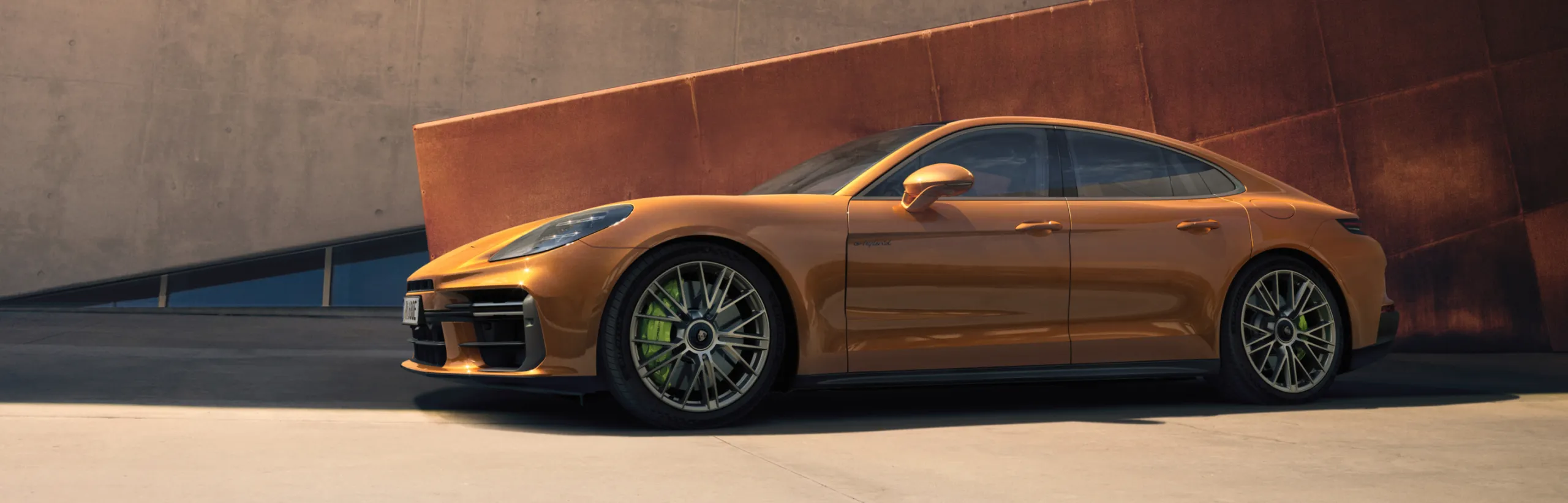 Porsche Panamera Turbo E-Hybrid i Madeira Gold Metallic står framför en betongvägg. Vy mot sidan.