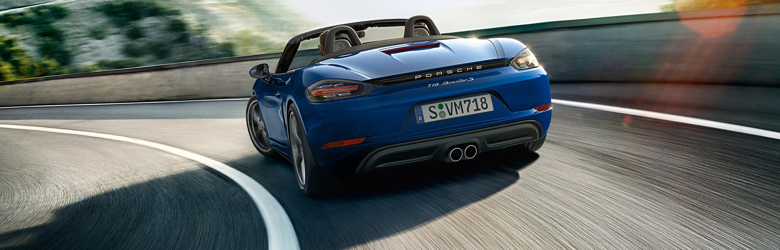 Porsche 718 Boxster S en color Azul Gentian Metalizado tomando una curva en una carretera de montaña. Vista de la parte trasera.
