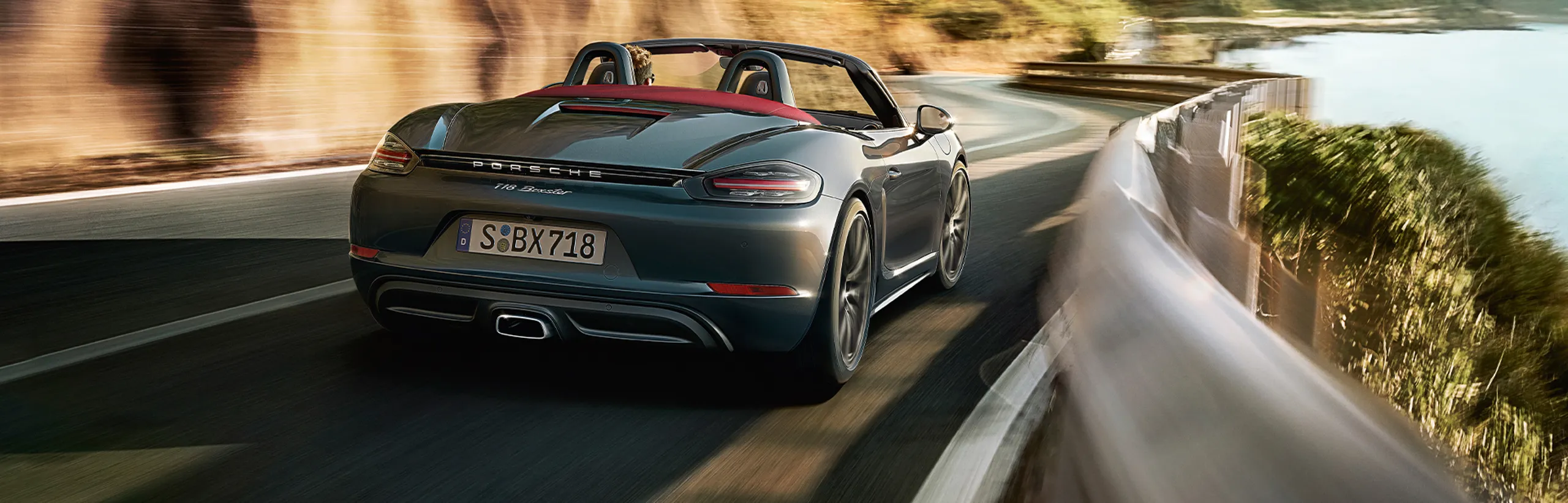 Porsche 718 Boxster in Agate Grey Metallic driving along a winding coastal road. View of the rear end.