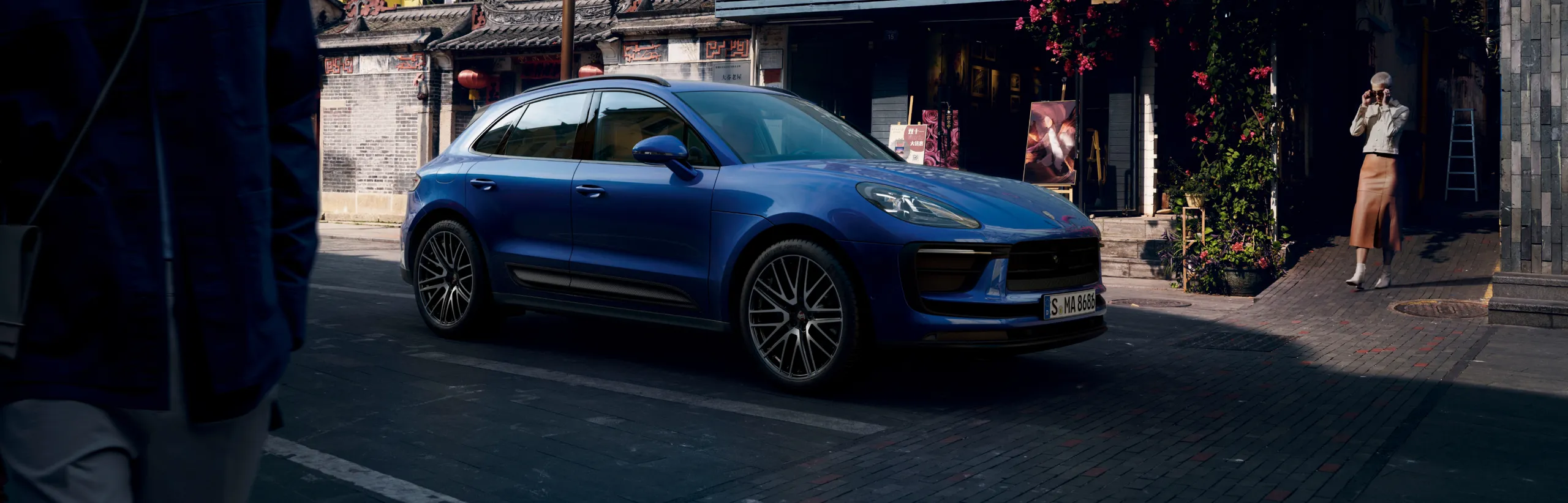 Porsche Macan em Azul Gentian metálico em uma rua em frente a uma galeria. Vista lateral.