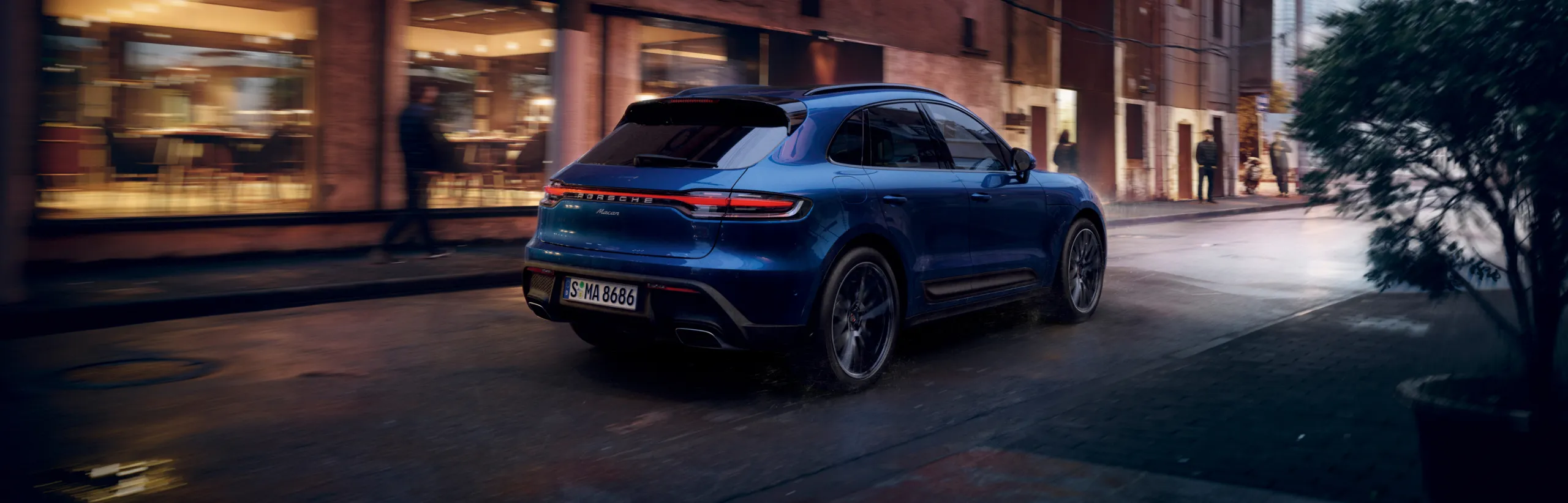 Porsche Macan цвета синий металлик (Gentian Blue Metallic) едет по городу. Вид сбоку и сзади.
