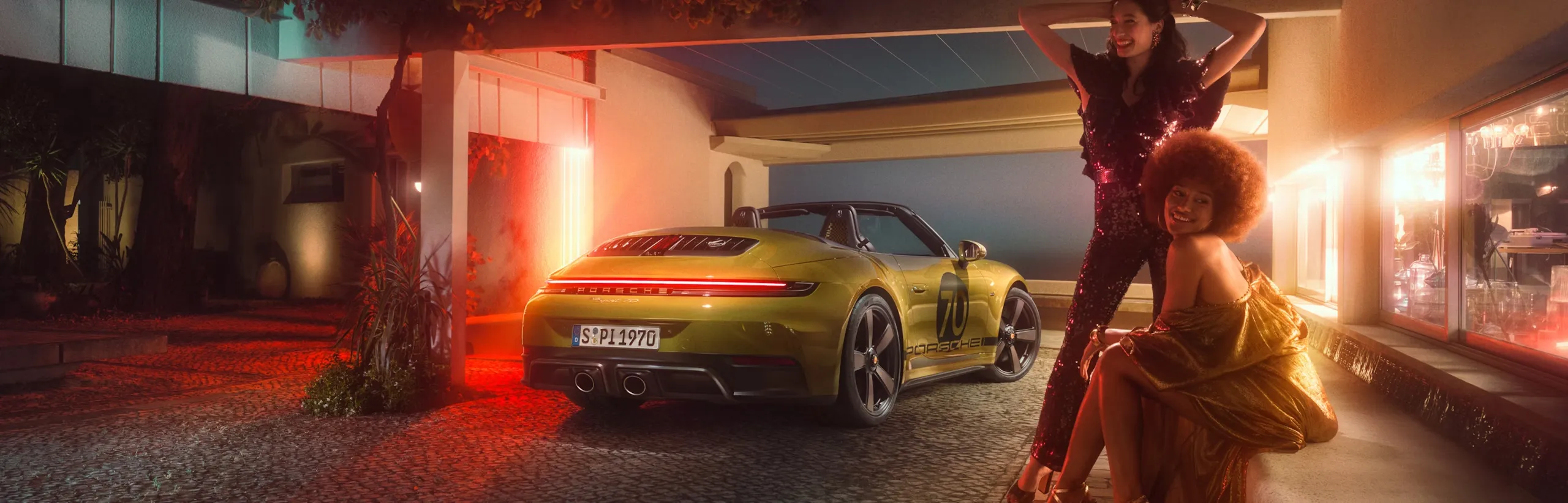Porsche 911 Spirit 70 in an Olive Neo paint finish parked under a carport at night. Rear and side view from behind. Two women wearing 70s clothing are posing in the foreground.