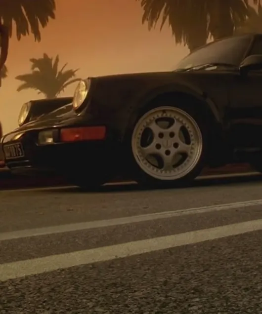 Zicht op de zijkant van een zwarte Porsche 911 Turbo 3.6 van de generatie 964 met op de achtergrond een zonsondergang en palmbomen. 