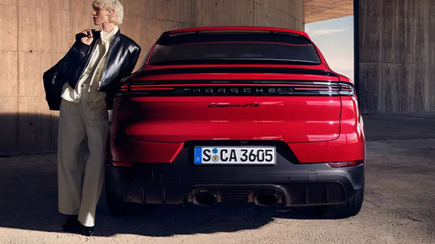 A woman with short, light-coloured hair and wearing a black leather jacket and brown leather pants poses next to a red Porsche Cayenne GTS. The scene is a parking garage with red accent lights.