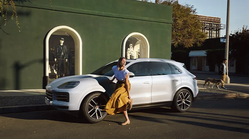 A woman in a white shirt and brown skirt gets out of a white Porsche Cayenne parked on a city street. A dark green building with two large windows displaying mannequins is in the background.
