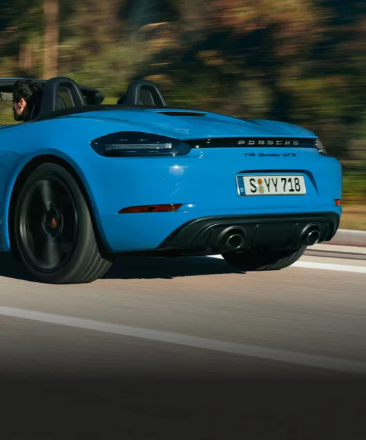 Porsche 718 Boxster GTS 4.0 en color Azul Shark circulando por una carretera. Vista de la parte trasera con ópticas traseras oscurecidas.
