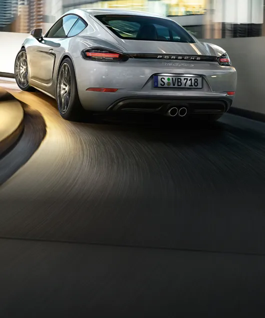 Porsche 718 Cayman S in GT Silver Metallic driving through a car park. Rear and side view.